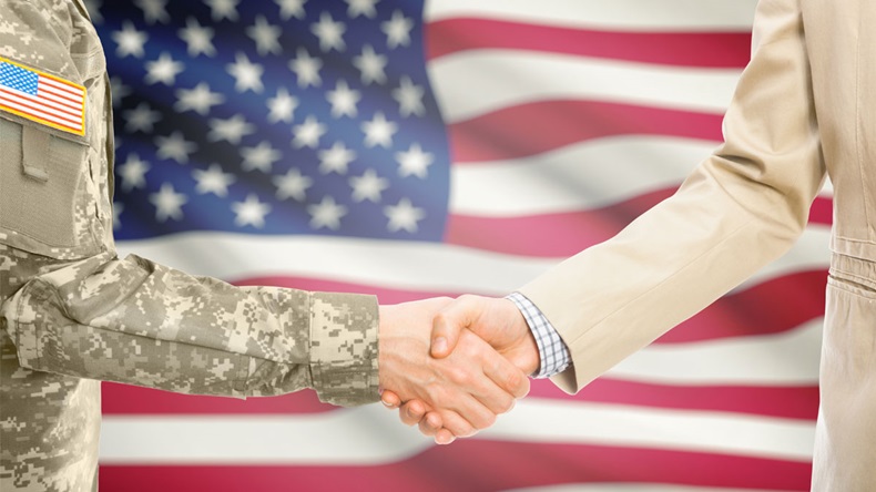 American soldier in uniform and civil man in suit shaking hands with national flag on background - United States