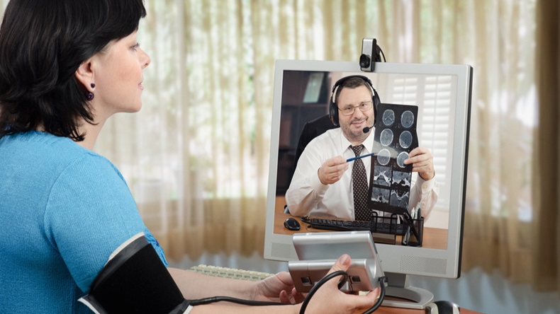 Woman has just measured her blood pressure and shows blood pressure gauges figures to virtual doctor. Male physician in headset is carefully looks it and explains brain x ray picture in the monitor - Image 