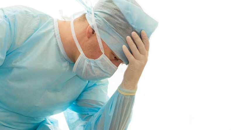 Upset surgeon in sterile gown and surgical mask props his head with hand