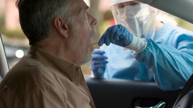 A medical technician in full protective gear collects a sample from a mature man sitting inside his car as part of the operations of a coronavirus mobile testing unit.