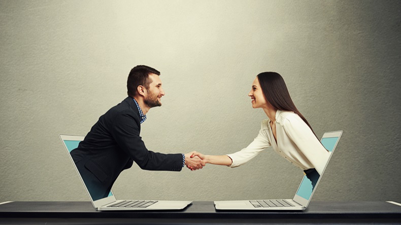 smiley businessman and businesswoman come out from laptop, shaking hands and looking at each other over dark grey background