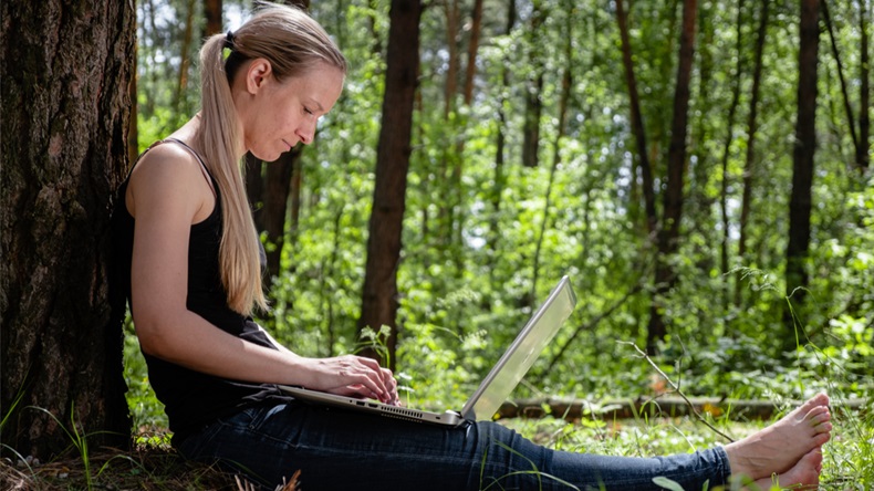 girl in the forest sits leaning against a tree, working on a laptop. rest at nature. in the background blurred silhouettes of trees. digital detox. Mental health