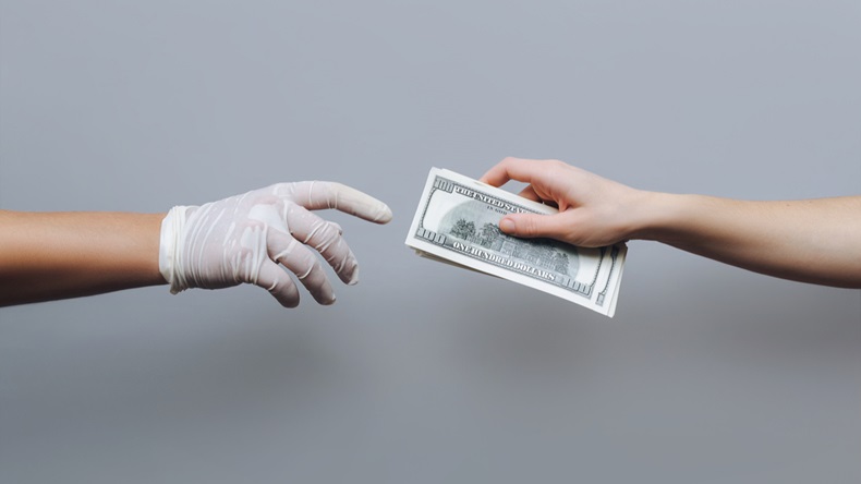 Hand of a woman giving money in the form of hundred-dollar bills to the doctor in a white medical glove. 