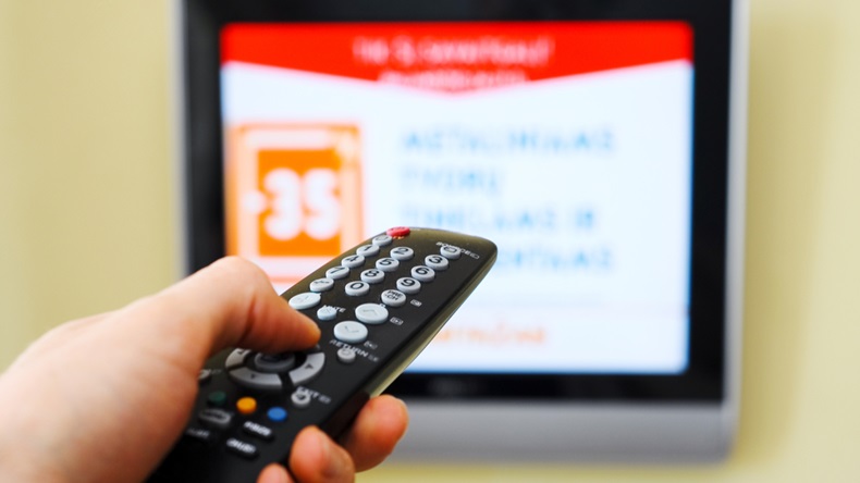 A TV screen is seen in the background, with a hand holding a remote in the foreground.
