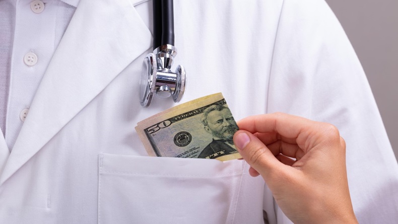 A hand slips a folded $50 bill into the pocket of a physician's white coat.