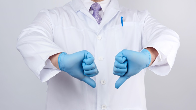 Doctor in a white coat with buttons shows a gesture of dislike with his hands, wearing blue medical gloves.
