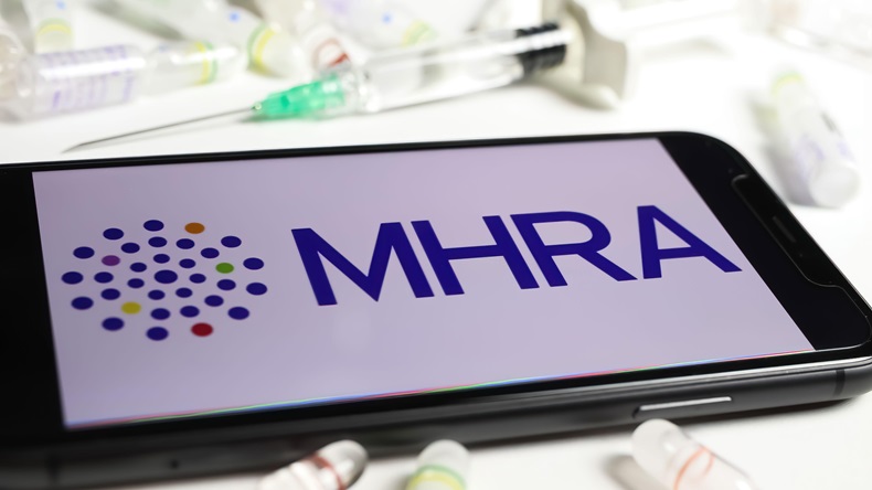 Closeup of mobile phone screen with logo lettering of UK MHRA agency, serum vials and syringe background.