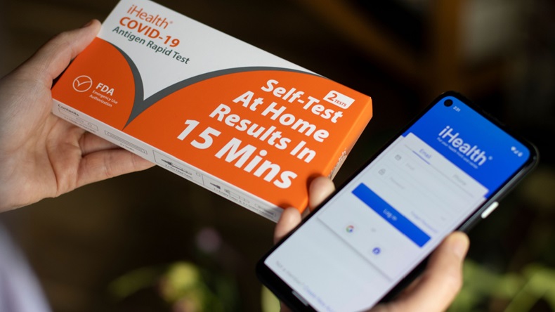 A woman's hands holding a free iHealth COVID-19 Antigen rapid test kit ordered from USPS website and a smartphone showing the iHealth COVID-19 Test app login page.