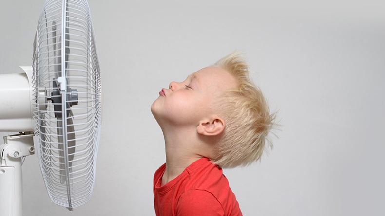 Blond boy tilts his head back in front of a fan, seemingly enjoying the cool air.