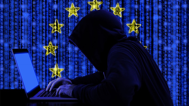 Hacker in a dark hoody sitting in front of a notebook with digital european flag and binary streams background cybersecurity concept