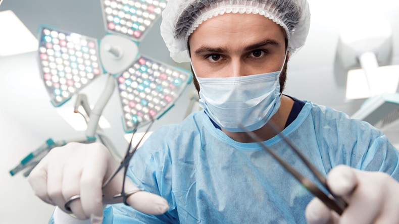 Nothing to be scared of! Professional male surgeon wearing protective uniform looking at the camera with surgical instruments in his hands surgical lamp on the background copyspace doctor medical job